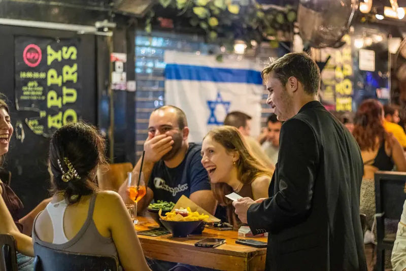 דני בוריסובר בקלוז-אפ ליד שולחן מלא אורחים צוחקים